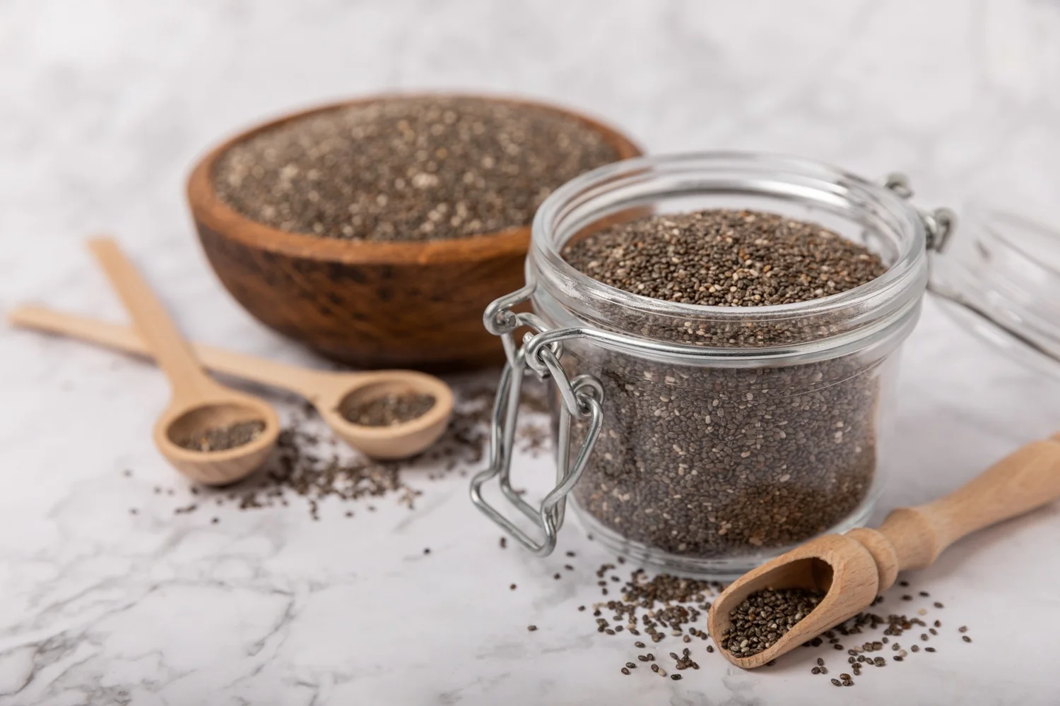 Glass jar and bowl of chia seeds with wooden spoons full of chia seeds fibre.